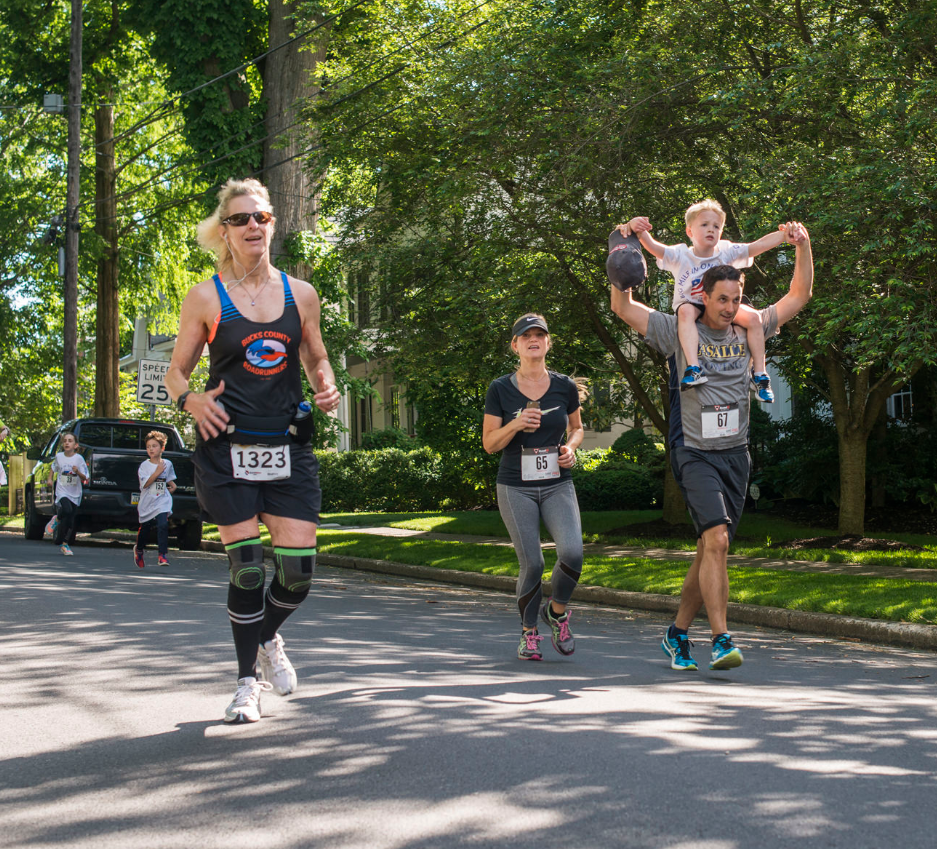 A woman in a Bucks County Roadrunners singlet wearing bib 1323 leading a group of runners down a sunny tree-lined residential street, with a dad carrying a young child on his shoulders and kids running behind her